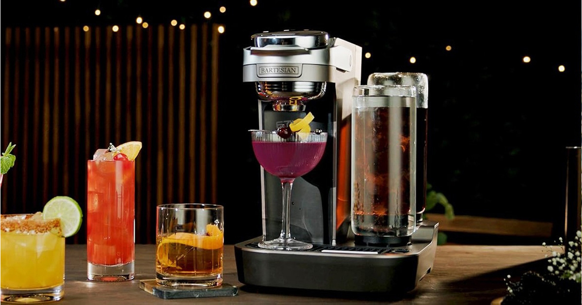 Bartesian Duet cordless cocktail maker preparing a purple cocktail, surrounded by colorful drinks including a margarita, a red cocktail and whiskey on a wooden bar under warm ambient lighting.