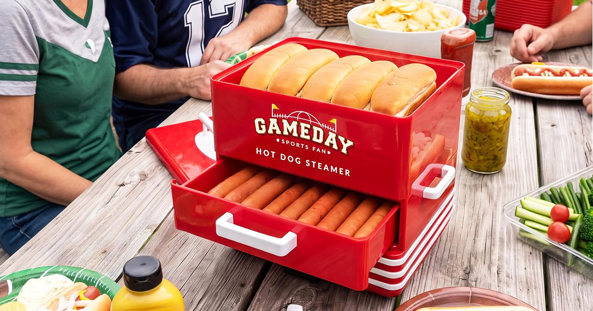 Red Nostalgia GameDay hot dog steamer and bun warmer filled with hot dogs and buns, displayed on an outdoor party table surrounded by family and friends wearing sports jerseys, with bowls of chips, condiments, pickles and fresh vegetables ready for game day.