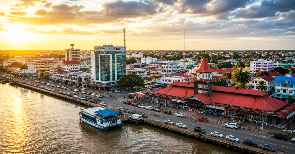Aerial sunset view of Georgetown, Guyana featuring the historic Stabroek Market clock tower, waterfront promenade, busy harbor, colonial architecture, and the vibrant capital city skyline along the Demerara River.
