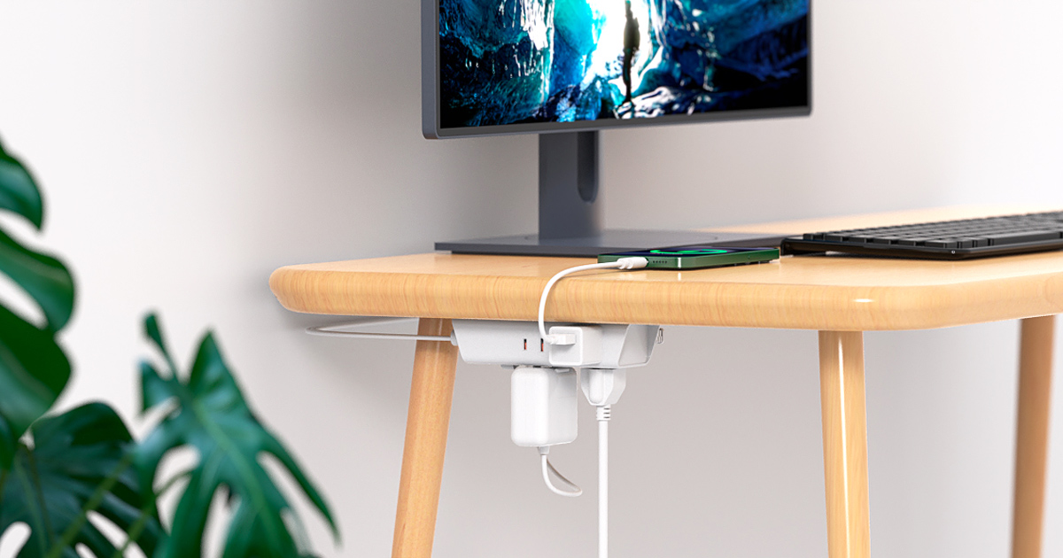 Flat plug power strip mounted under a desk charging devices, with a smartphone on a wooden workstation beside a computer monitor.