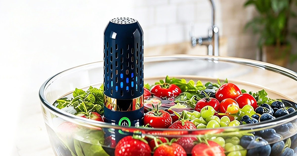 A dark blue fruit and vegetable purifier device submerged in a glass bowl of water, cleaning fresh strawberries, tomatoes, grapes, and blueberries in a kitchen setting.
