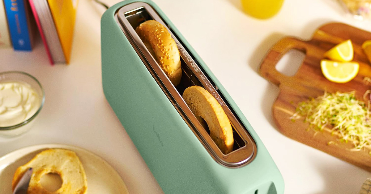 Mint-green Bella Fits-Anywhere 2-Slice Slim Toaster with a long slot holding toasted bagel slices, surrounded by a plate of bagels, cream cheese, lemon wedges, and fresh ingredients on a bright kitchen countertop.