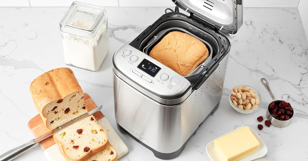 A stainless steel bread maker open on a white marble countertop with a freshly baked loaf inside, alongside a sliced loaf with fruit and nuts on a wooden cutting board, butter, and baking ingredients.