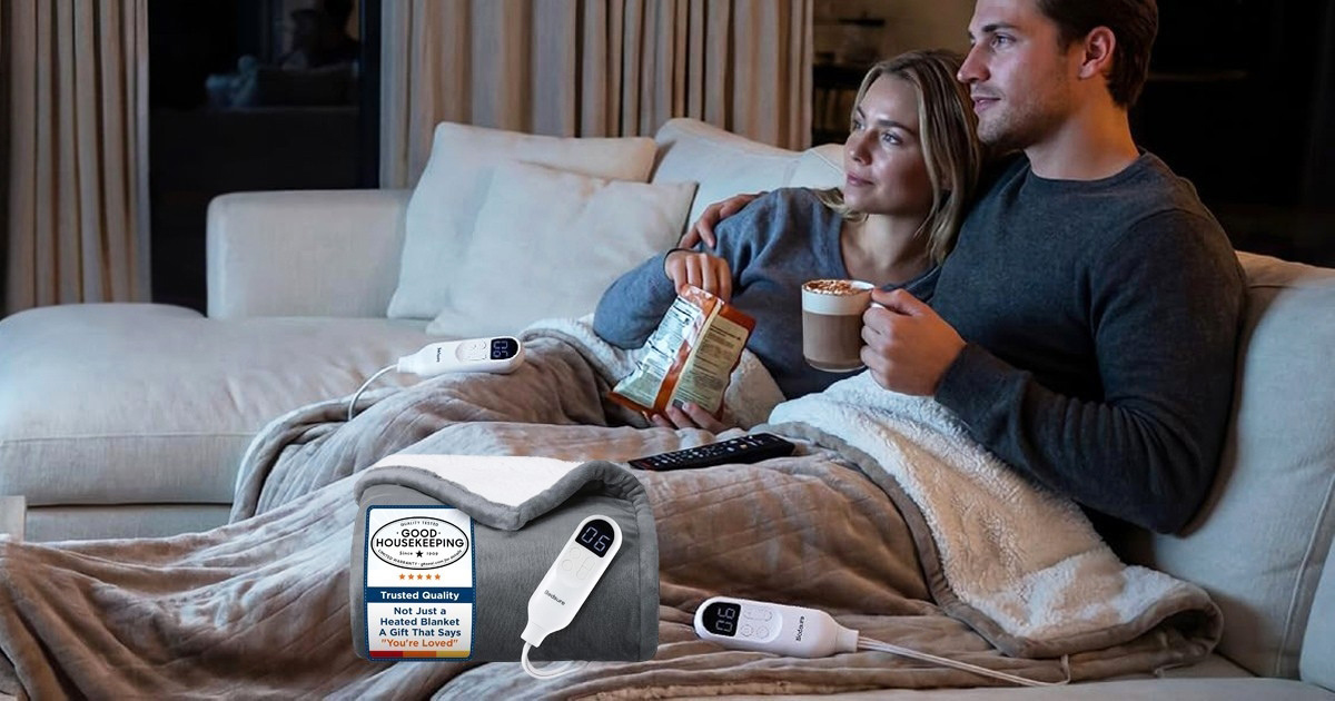 A couple relaxes together on a sofa under a soft gray Bedsure heated throw blanket, holding hot drinks and snacks while watching television. The blanket's temperature controllers and a Good Housekeeping Seal are visible in the foreground, highlighting its cozy design and product recognition.