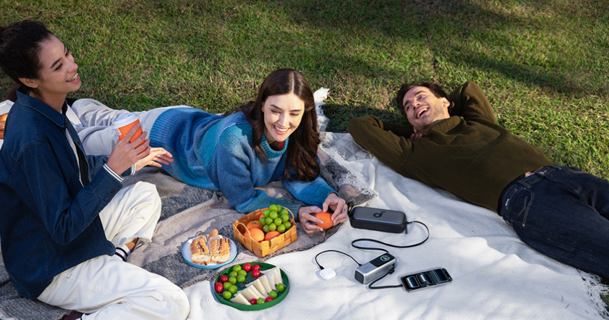 Three friends on a picnic using a portable power gadget to keep their devices charged outdoors.